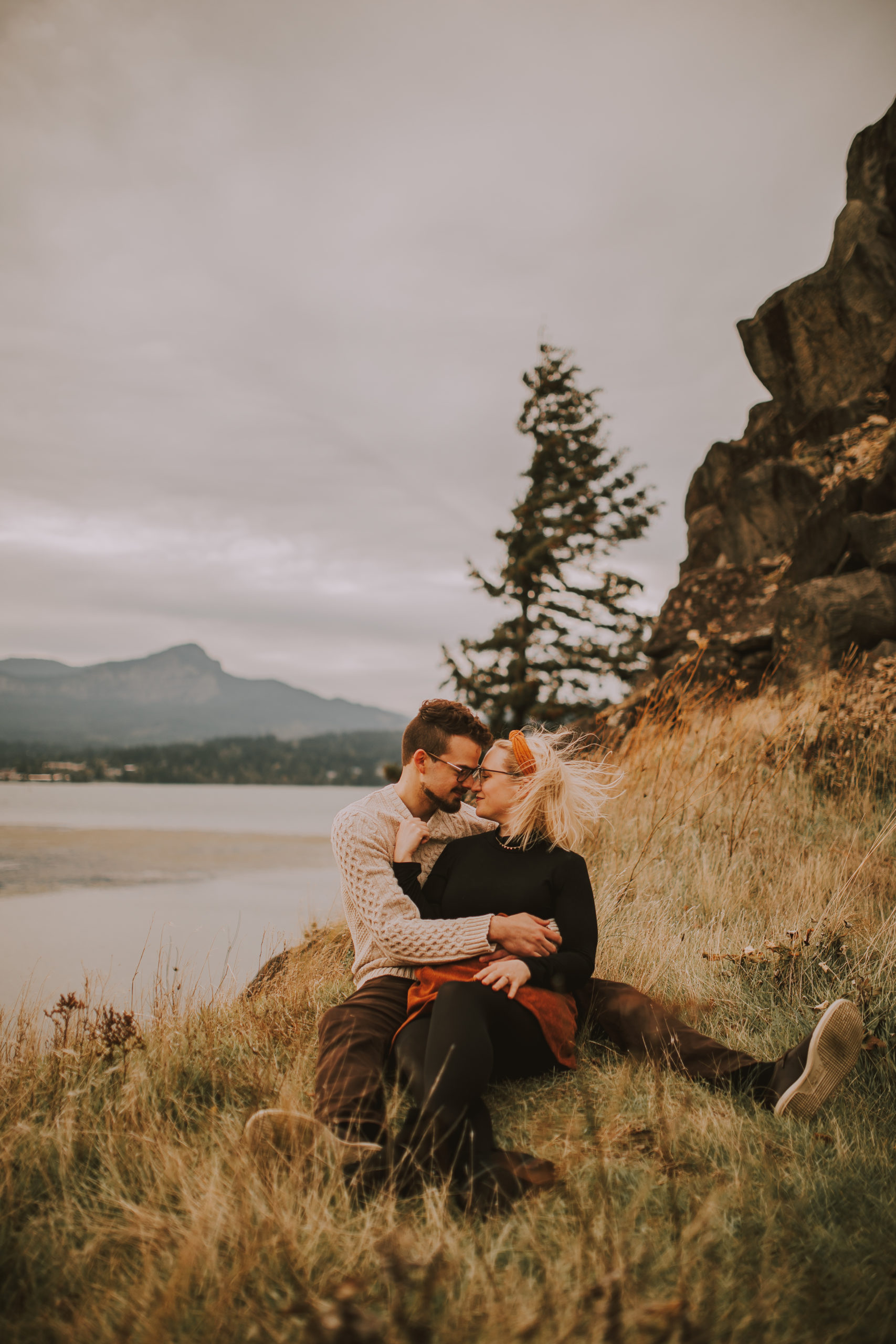 engaged couple laying on the floor cliffside in front of mountains and lake; leaning in for an intimate kiss