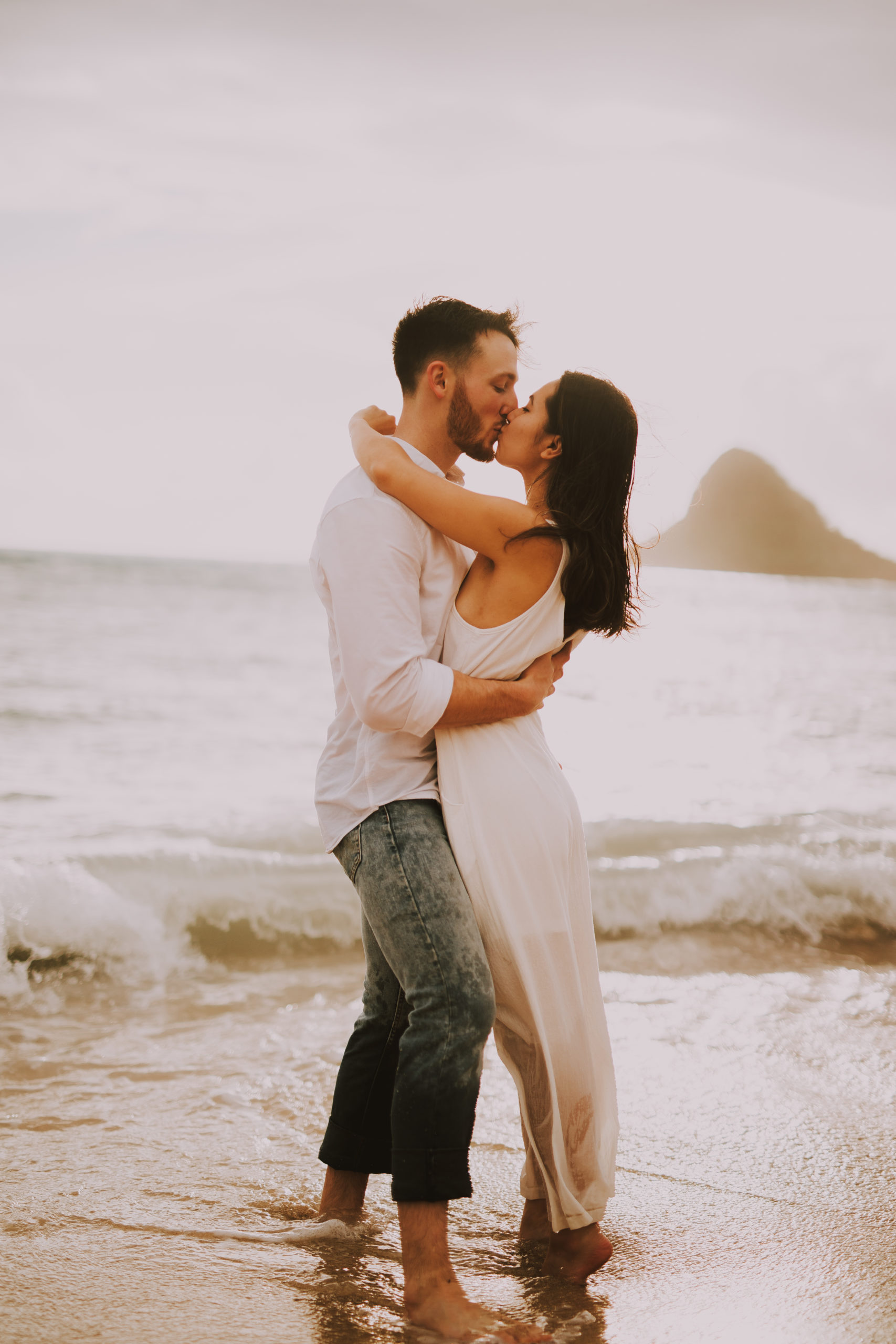 Bride and groom kiss on the beach as waves crash onto the shore in Honolulu, Hawaii Kualoa Ranch