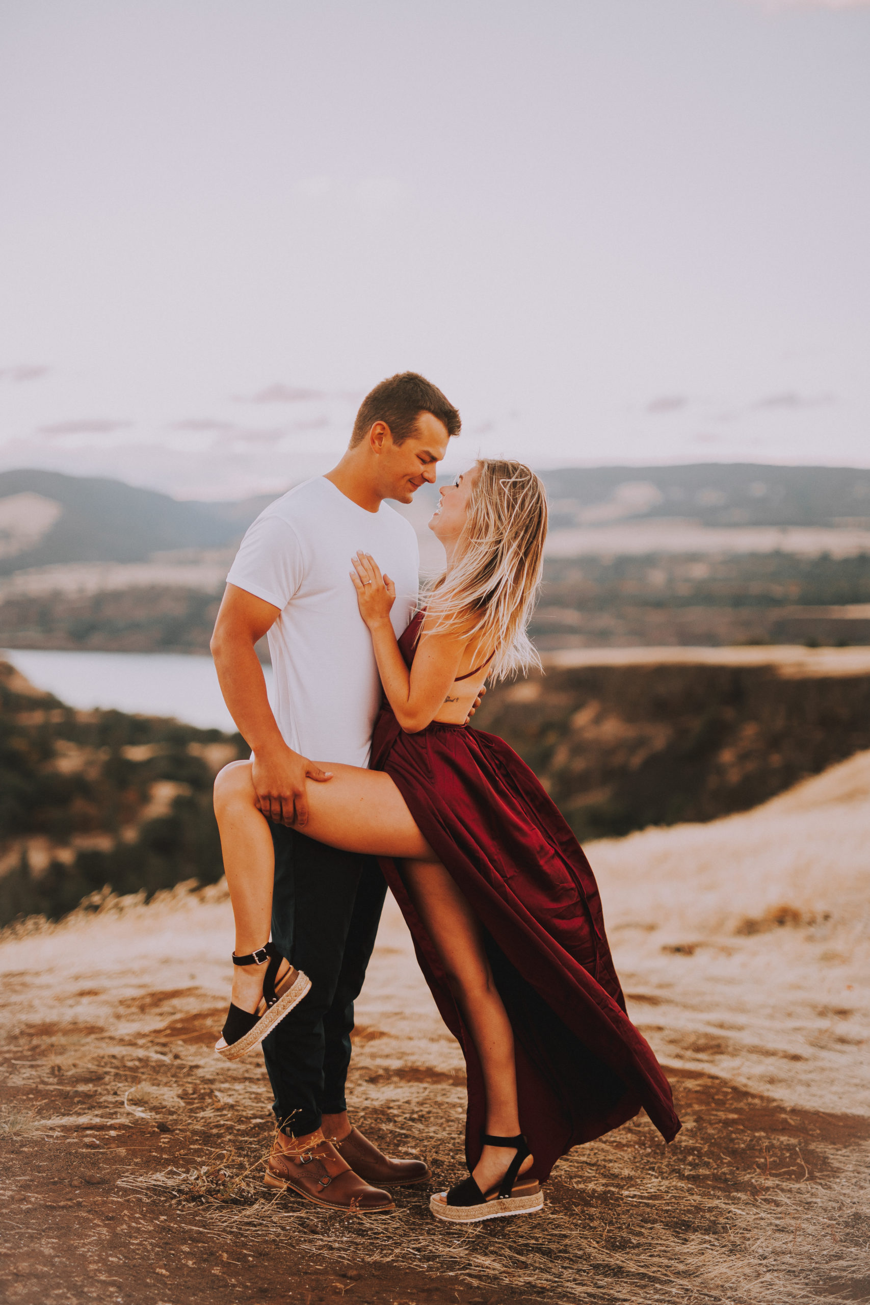 Engaged couple lean in for a kiss with the grooms hand holding the brides leg at Rowena Crest Viewpoint in Wasco County, Oregon in front of beautiful mountains