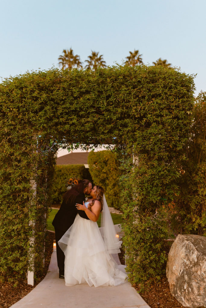 The groom leans in to kiss his bride under a beautiful arch of greenery at the Gather Estate wedding venue in Mesa Arizona