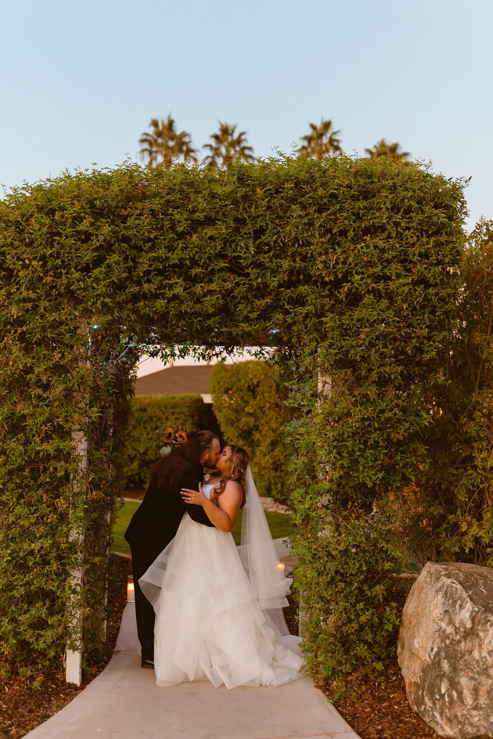 The bride leans in to kiss his bride underneath a arch filled with greenery